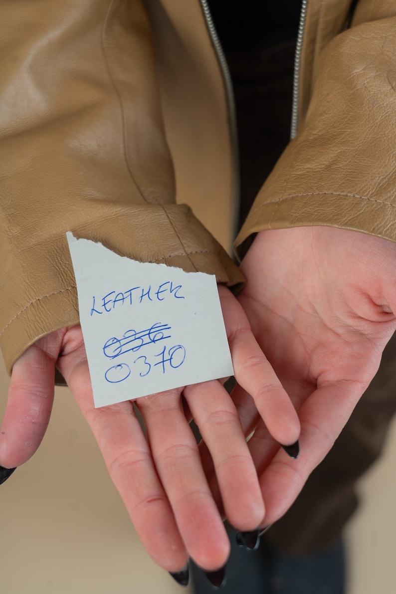 Pode incluir: Um close-up de uma jaqueta de couro marrom com uma nota escrita &agrave; m&atilde;o que diz "LEATHER" e tem os n&uacute;meros "036" e "0370" escritos nela.