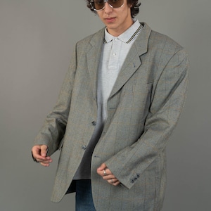 May include: A gray plaid blazer with a single button closure. The blazer has a notched lapel and two flap pockets. The model is wearing a gray polo shirt and blue jeans.