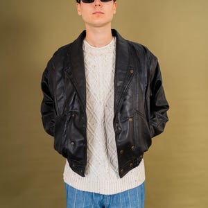 May include: A person wearing a black leather jacket with a button-up front, over a white sweater and blue striped pants. The person is also wearing black sunglasses. The background is a solid olive green.