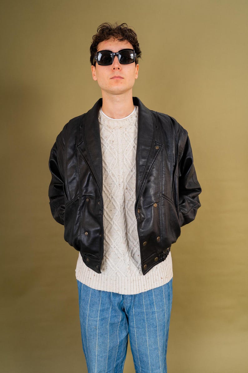 May include: A person wearing a black leather jacket with a button-up front, over a white sweater and blue striped pants. The person is also wearing black sunglasses. The background is a solid olive green.