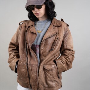 May include: A brown leather jacket with a zip closure and a zipped pocket on the left side. The jacket has a grey shirt underneath and a grey baseball cap with the letters "NY" on it.