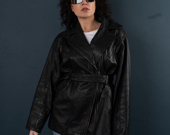 May include: A woman wearing a black leather jacket with a belt, blue jeans, and sunglasses. The jacket has a zipper closure and a collar.
