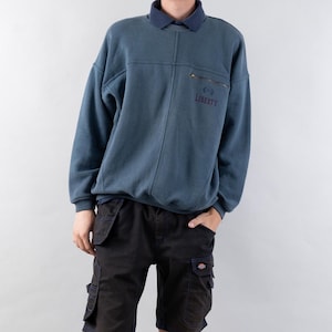 Puede incluir: Una sudadera azul con un bolsillo con cremallera y la palabra "LIBERTY" bordada en el bolsillo.