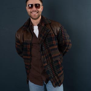 Könnte beinhalten: Ein Mann trägt eine braune und schwarze karierte Jacke mit Ledereinsätzen. Er trägt ein braunes Hemd mit Knöpfen und eine blaue Jeans. Er lächelt und trägt eine Sonnenbrille.