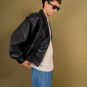 May include: A black leather jacket with a double-breasted design. The jacket is worn over a cream-colored sweater and paired with blue striped pants. The person in the image is wearing black sunglasses.