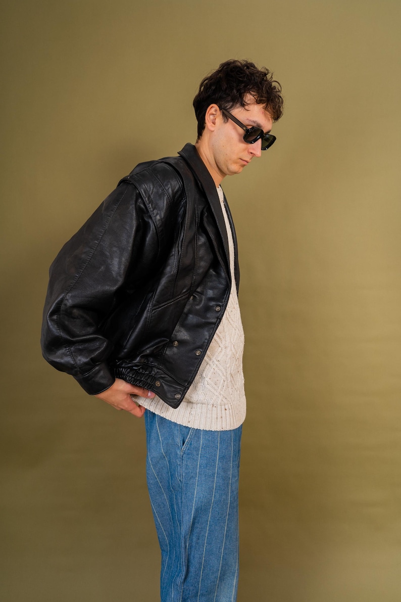 May include: A black leather jacket with a double-breasted design. The jacket is worn over a cream-colored sweater and paired with blue striped pants. The person in the image is wearing black sunglasses.
