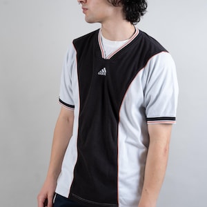 Op de afbeelding: Een zwart-wit T-shirt met korte mouwen en een contrasterend kleurenpaneelontwerp. Het shirt heeft een V-hals en het Adidas-logo op de voorkant.