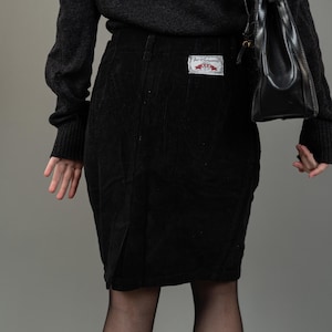 Könnte beinhalten: Ein schwarzer Cordrock mit einem roten und weißen Etikett, auf dem "Jet Company" steht. Der Rock wird mit einem schwarzen Pullover und schwarzen Strumpfhosen getragen.