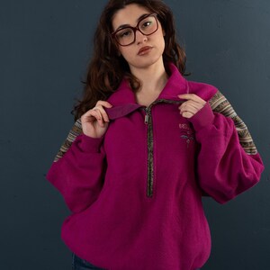 May include: A magenta fleece pullover with a half-zip closure and brown trim. The sleeves have a brown and white geometric pattern. The pullover has a small embroidered logo on the left chest.