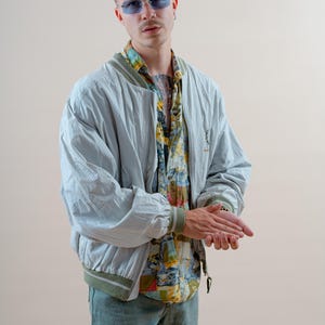May include: A light blue bomber jacket with a green trim, paired with a colorful patterned shirt and blue jeans. The jacket has a logo on the chest. The person is wearing blue tinted glasses.