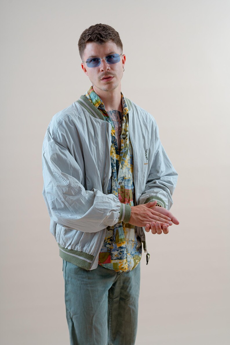 May include: A light blue bomber jacket with a green trim, paired with a colorful patterned shirt and blue jeans. The jacket has a logo on the chest. The person is wearing blue tinted glasses.