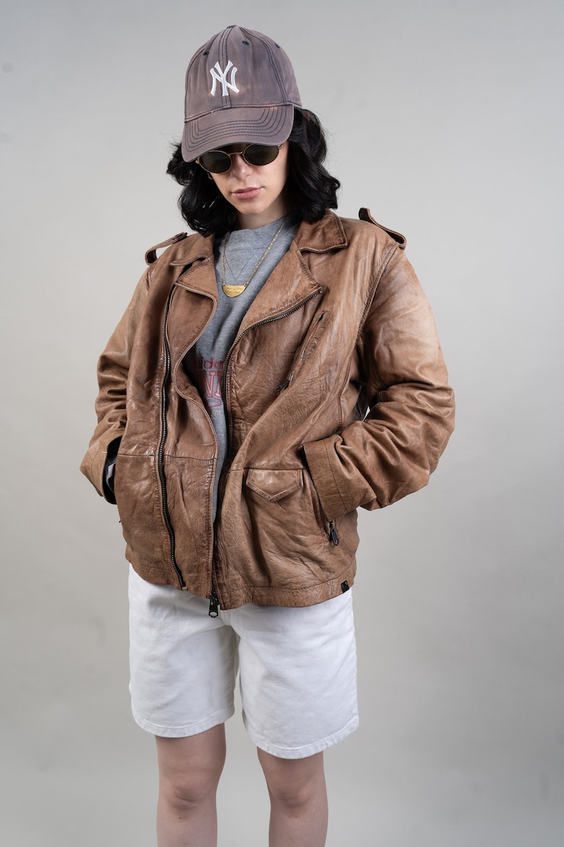 May include: A brown leather biker jacket with a zip fastening and multiple pockets. The jacket is worn over a grey t-shirt and white shorts. The person is wearing a brown baseball cap with a white "NY" logo.