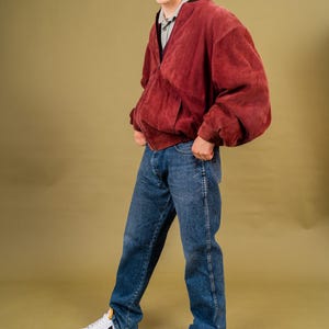 May include: A burgundy suede bomber jacket with a relaxed fit, paired with blue denim jeans and white sneakers. The jacket features a zip-up front and pockets. The sneakers have black accents. The outfit is set against a solid olive-green background.
