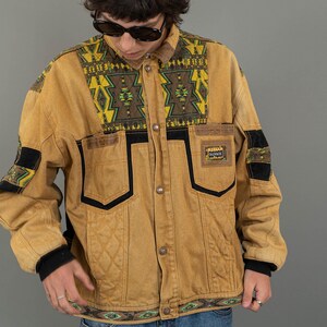 Puede incluir: Una chaqueta vaquera marrón con un patrón geométrico en el cuello y los bolsillos. La chaqueta tiene un ribete negro y un parche en el pecho izquierdo que dice "Rampage".