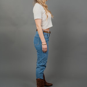 Peut inclure: Un haut court &agrave; manches courtes de couleur claire, associ&eacute; &agrave; un jean bleu clair taille haute et une ceinture en cuir marron. La tenue est compl&eacute;t&eacute;e par des bottes de cowboy en cuir marron. Le fond est gris uni.