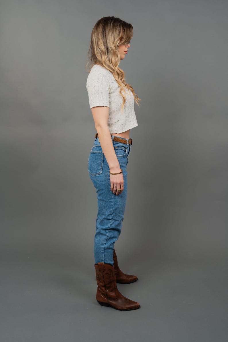 Peut inclure: Un haut court &agrave; manches courtes de couleur claire, associ&eacute; &agrave; un jean bleu clair taille haute et une ceinture en cuir marron. La tenue est compl&eacute;t&eacute;e par des bottes de cowboy en cuir marron. Le fond est gris uni.