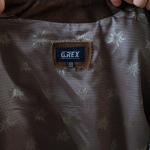Op de afbeelding: Close-up van de binnenkant van een bruine su&egrave;de jas. De voering is een bruine stof met een patroon met een herhalend sterrenpatroon. Een label leest "G.REX" met het nummer "50" eronder.