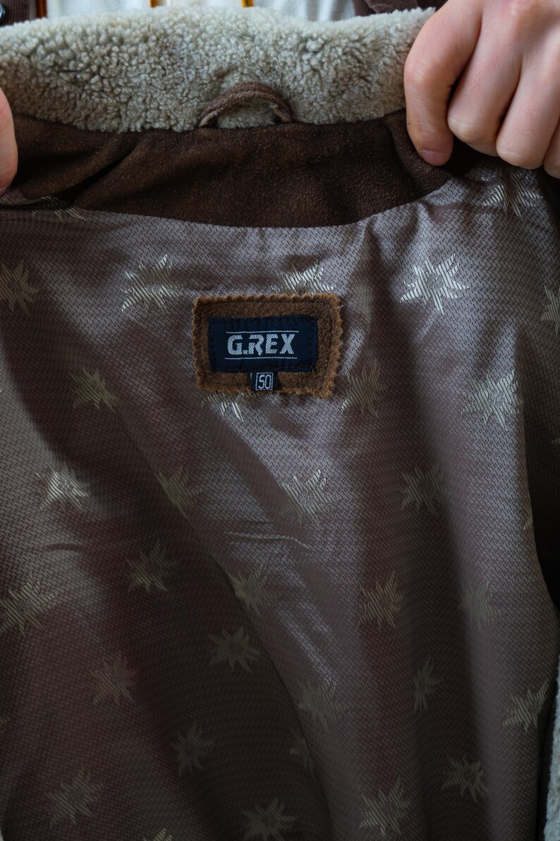 Op de afbeelding: Close-up van de binnenkant van een bruine su&egrave;de jas. De voering is een bruine stof met een patroon met een herhalend sterrenpatroon. Een label leest "G.REX" met het nummer "50" eronder.