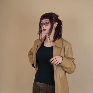 Pode incluir: Uma mulher vestindo uma jaqueta de couro marrom sobre uma blusa preta e cal&ccedil;as de veludo cotel&ecirc; marrom. Ela est&aacute; usando &oacute;culos de sol e tem cabelo roxo escuro.