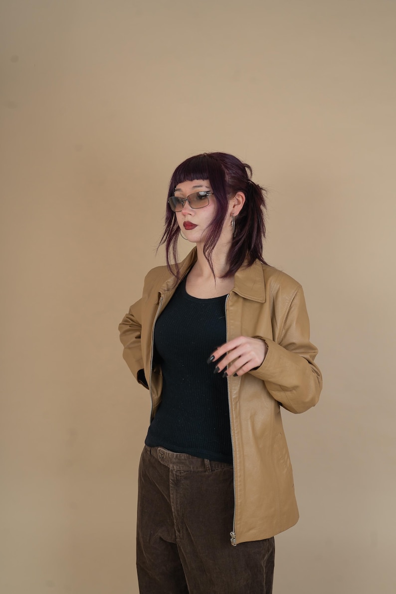 Pode incluir: Uma mulher vestindo uma jaqueta de couro marrom sobre uma blusa preta e cal&ccedil;as de veludo cotel&ecirc; marrom. Ela est&aacute; usando &oacute;culos de sol e tem cabelo roxo escuro.
