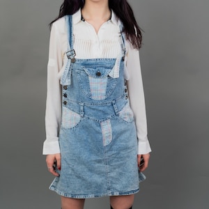 Könnte beinhalten: Ein hellblaues Jeans-Overallkleid mit einem weißen Hemd mit Knöpfen darunter. Das Kleid hat ein Karomuster auf den Taschen und einen hellblauen Jeansrock.
