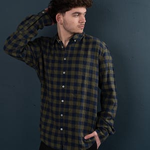 Peut inclure: Un homme portant une chemise à manches longues à carreaux verts et bleus avec un col boutonné. Il se tient devant un mur bleu foncé.