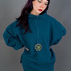Könnte beinhalten: Türkisfarbener Fleece-Pullover mit einem Viertel-Reißverschluss am Hals und einem runden, gestickten Aufnäher. Der Aufnäher zeigt ein geometrisches Design mit gelben, blauen und grünen Akzenten. Der Pullover hat lange Ärmel und eine lockere Passform.