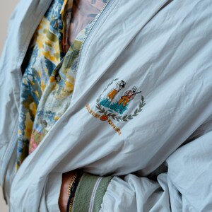 May include: A light gray zip-up jacket with a green ribbed collar and cuffs featuring white stripes. The chest displays an embroidered emblem of two figures and the text BANANA REPUBLIC. Underneath, a yellow, blue, and white patterned shirt is visible.