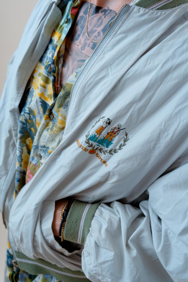 May include: A light gray zip-up jacket with a green ribbed collar and cuffs featuring white stripes. The chest displays an embroidered emblem of two figures and the text BANANA REPUBLIC. Underneath, a yellow, blue, and white patterned shirt is visible.