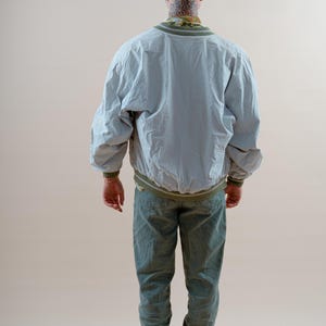 May include: A light gray bomber jacket with olive green ribbed trim at the collar, cuffs, and hem, paired with olive green trousers and black and white sneakers. The jacket features a loose fit and long sleeves, worn over a patterned shirt.