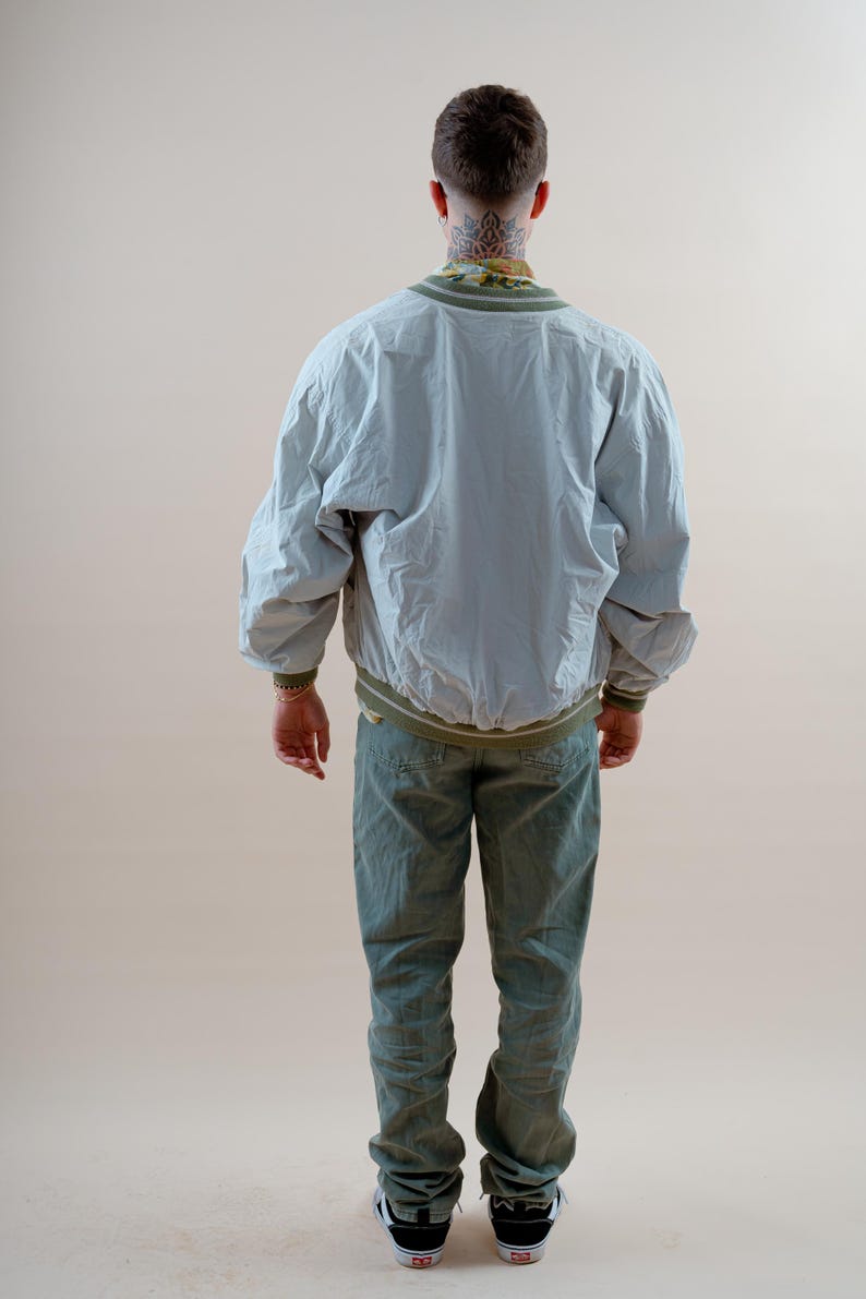 May include: A light gray bomber jacket with olive green ribbed trim at the collar, cuffs, and hem, paired with olive green trousers and black and white sneakers. The jacket features a loose fit and long sleeves, worn over a patterned shirt.