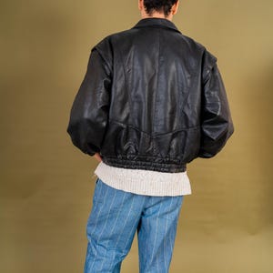 May include: Black leather bomber jacket with a ribbed hem and a collared neckline. The jacket is paired with blue and white striped pants and a white sweater. The model is facing away from the camera.