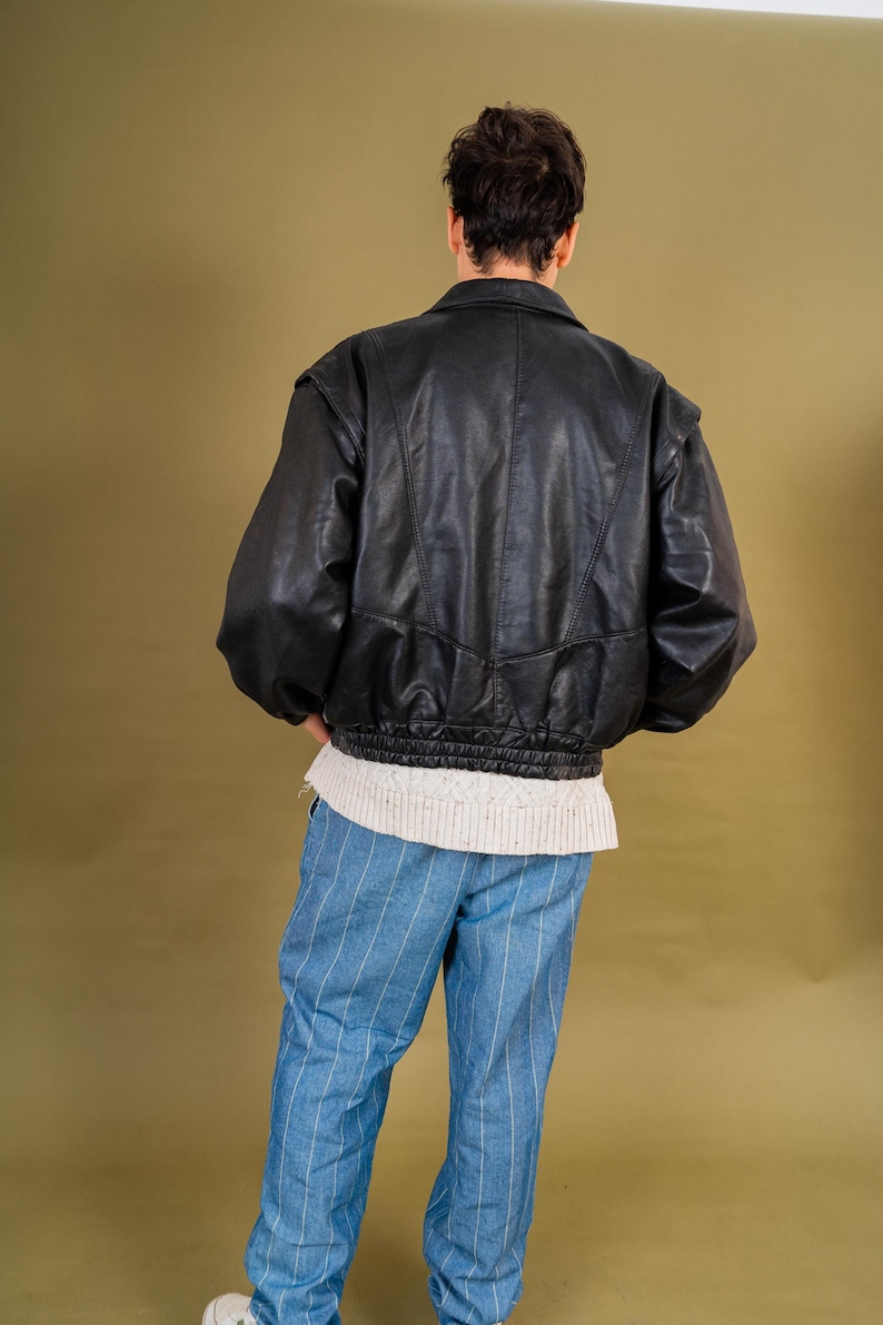 May include: Black leather bomber jacket with a ribbed hem and a collared neckline. The jacket is paired with blue and white striped pants and a white sweater. The model is facing away from the camera.
