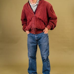 May include: A person wearing a burgundy zip-up jacket, blue jeans, and white sneakers with black accents. The jacket has a V-neck and is layered over a collared shirt. The outfit is casual and stylish.