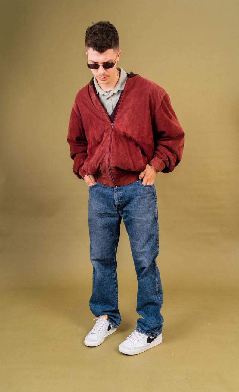 May include: A person wearing a burgundy zip-up jacket, blue jeans, and white sneakers with black accents. The jacket has a V-neck and is layered over a collared shirt. The outfit is casual and stylish.
