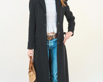 Puede incluir: Un abrigo largo gris oscuro con cierre de botones, usado sobre una parte superior blanca y jeans azules. El abrigo llega hasta la rodilla y tiene un diseño clásico. Se sostiene un bolso marrón en la mano. El atuendo se completa con botas marrones.