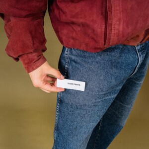 May include: A person wearing a burgundy jacket and blue jeans. The person is holding a small white tag that reads "18259 PANTS". Black sunglasses are held in the other hand.