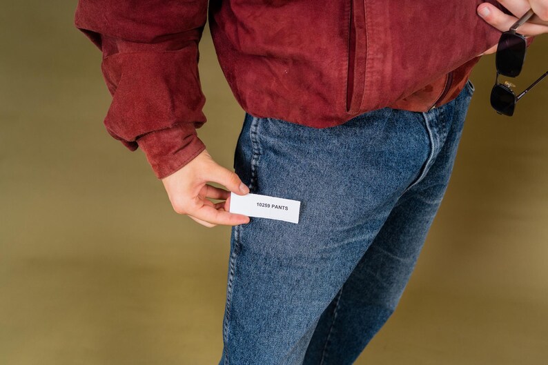 May include: A person wearing a burgundy jacket and blue jeans. The person is holding a small white tag that reads "18259 PANTS". Black sunglasses are held in the other hand.