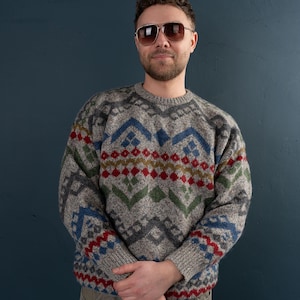 May include: A man wearing a grey sweater with a colourful geometric pattern. The sweater has blue, red, green and yellow accents. He is wearing sunglasses and has a beard.