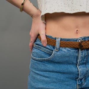 Peut inclure: Un bracelet jonc dor&eacute; sur un poignet, associ&eacute; &agrave; un pull court blanc mouchet&eacute; et un jean bleu clair. Une ceinture tress&eacute;e marron avec une boucle en m&eacute;tal accessoirise le jean. L'ensemble est pos&eacute; sur un fond gris.