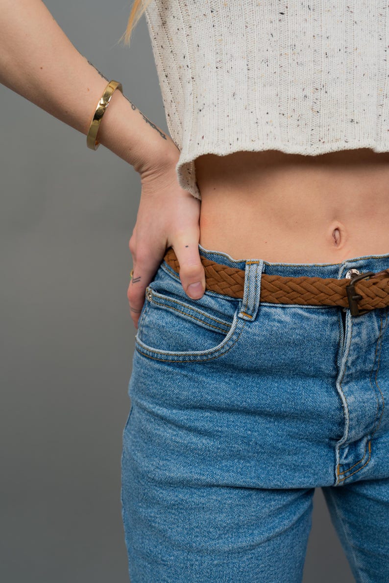 Peut inclure: Un bracelet jonc dor&eacute; sur un poignet, associ&eacute; &agrave; un pull court blanc mouchet&eacute; et un jean bleu clair. Une ceinture tress&eacute;e marron avec une boucle en m&eacute;tal accessoirise le jean. L'ensemble est pos&eacute; sur un fond gris.