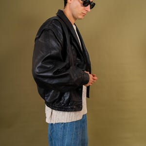May include: A black leather jacket with a bomber style, worn over a cream-colored sweater and blue pinstripe jeans. The jacket has a classic collar and a zipper closure. The person is wearing black sunglasses.