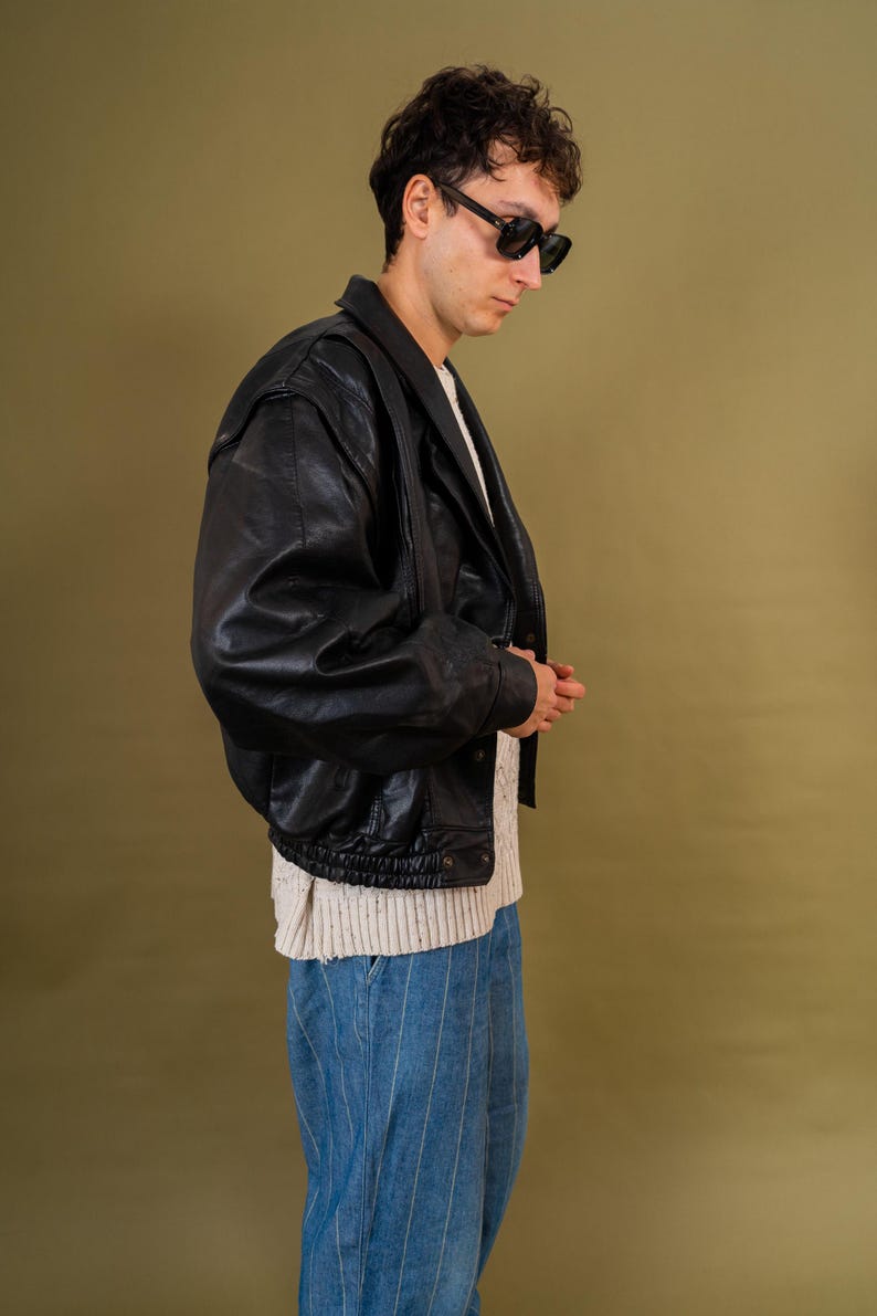 May include: A black leather jacket with a bomber style, worn over a cream-colored sweater and blue pinstripe jeans. The jacket has a classic collar and a zipper closure. The person is wearing black sunglasses.