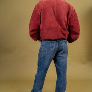 May include: A person wearing a burgundy suede jacket with a brown collar and blue jeans. The jeans are a medium wash denim. White sneakers complete the outfit. The background is a solid olive green.