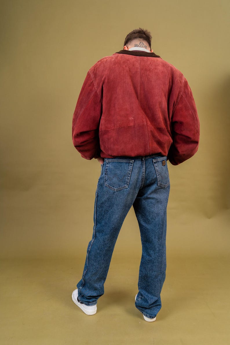May include: A person wearing a burgundy suede jacket with a brown collar and blue jeans. The jeans are a medium wash denim. White sneakers complete the outfit. The background is a solid olive green.