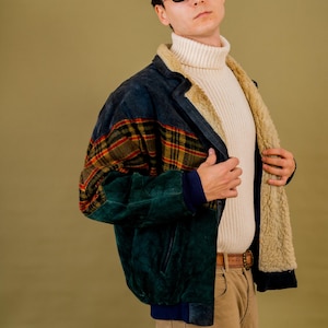 May include: A vintage suede jacket in shades of green and navy blue with a plaid pattern on the upper sleeves. The jacket has a cream-coloured shearling lining and is paired with a cream turtleneck and tan trousers.