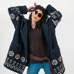 Op de afbeelding: Een vrouw draagt een donkerblauwe jas met witte en beige bloempatronen op de mouwen en zoom. De jas wordt gecombineerd met een bruine trui, een blauwe spijkerbroek en een bruine riem. Ze draagt ook een zonnebril.