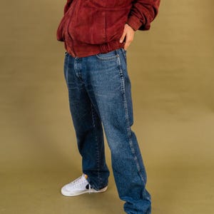 May include: A person wearing a burgundy suede jacket, blue denim jeans, and white sneakers with a black swoosh. The jeans have a classic five-pocket design and a relaxed fit. The outfit is set against a solid olive-green background.