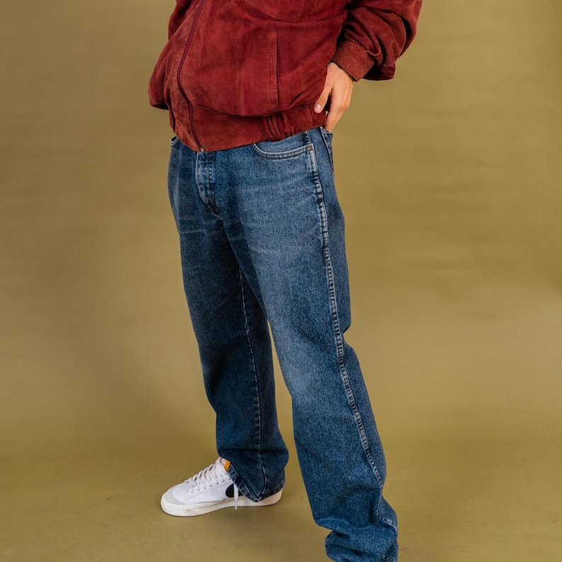 May include: A person wearing a burgundy suede jacket, blue denim jeans, and white sneakers with a black swoosh. The jeans have a classic five-pocket design and a relaxed fit. The outfit is set against a solid olive-green background.