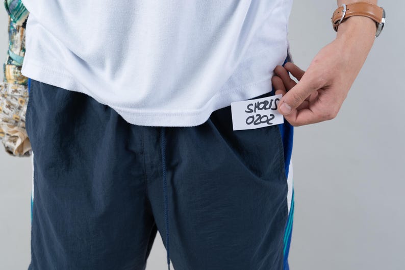 May include: A white t-shirt with a navy blue pair of shorts. A white tag is attached to the shorts with the text "SHORTS 0202" written in black marker.